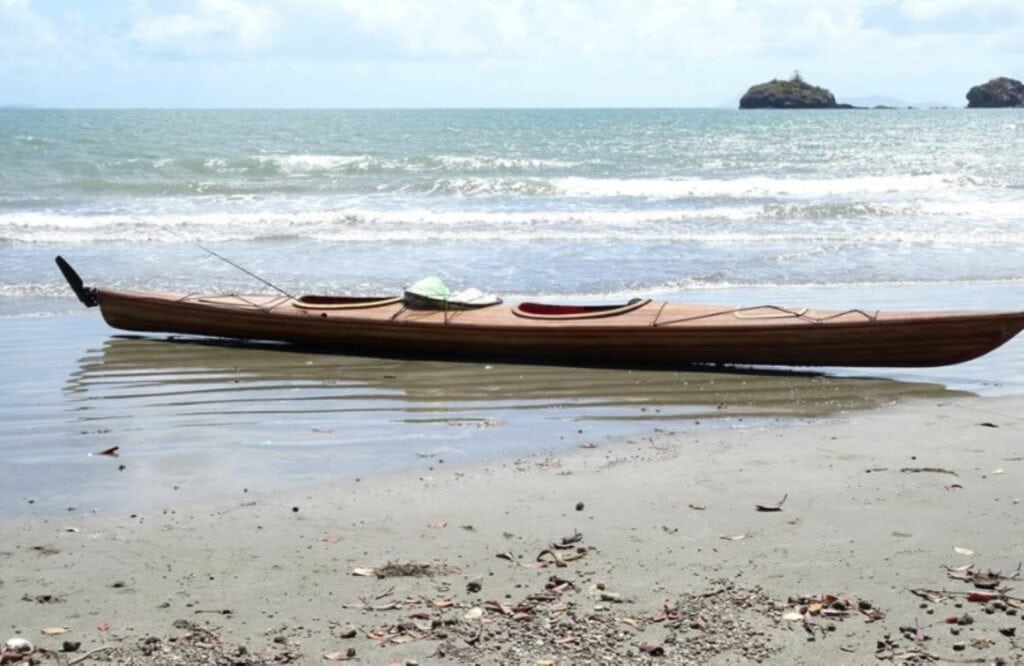 A strip planked double Kayak on a beach with a fishing rod hanging out the rear cockpit. 