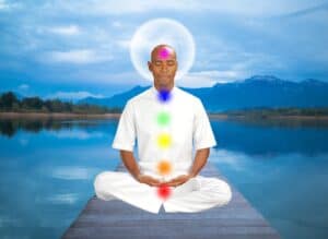 A photo of a guy in a yoga pose with a lake and mountain in the background, a full moon behind his head and coloured dots down his white shirt to illustrate the Reiki points. Used in the article, Does Reiki Healing Work or is it a Scam