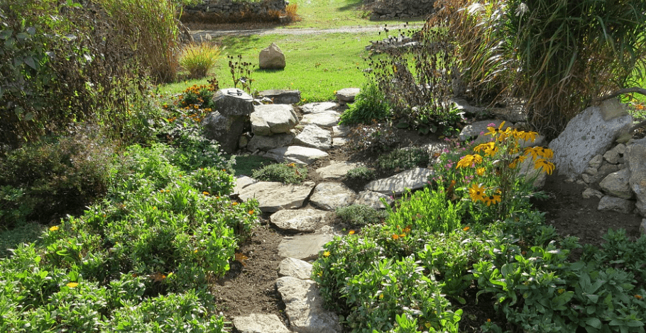 A rock garden path leading to a open grassed area. Each side of the path are bushy plants and flowers. Used in the article why are landscaping businesses unprofitable.