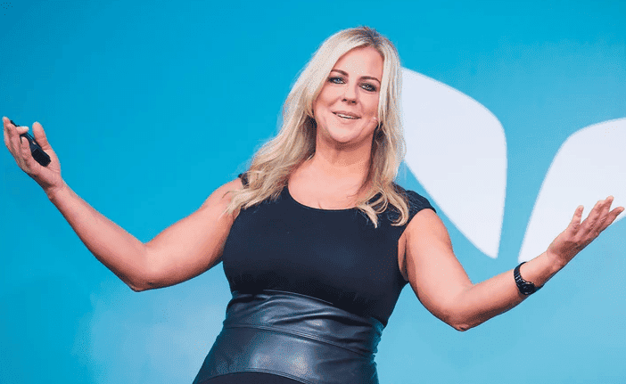 A photo of Christie Marie Sheldon in a black top and skirt with her arms outstretched, and blonde hair loose standing in front of a blue background with the white wings of mindvalley to the left of her. Used  in the article The Ultimate Mindvalley University Review