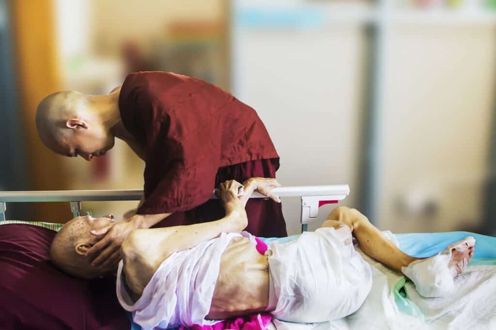 A monk offering comfort to a dying patient on a bed. Used in the article Is there a demand for death doulas?