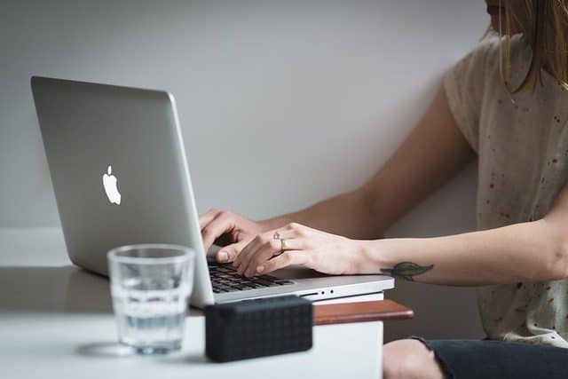 A below shoulder shot of a women sitting at a desk in a quiet corner working on an Apple computer with a glass of water nearby.  Used in the article: Online Education for stay-at-home parents. 