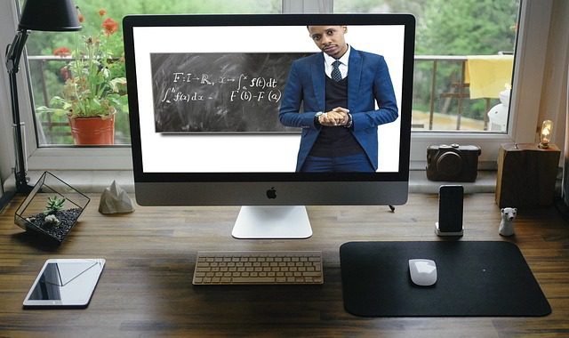 A large computer monitor on a table with an image of a teacher in front of a blackboard with some math formulas written on it. In the background a garden can be seen through a large window. Used in the article Online Education for stay-ay-home parents.